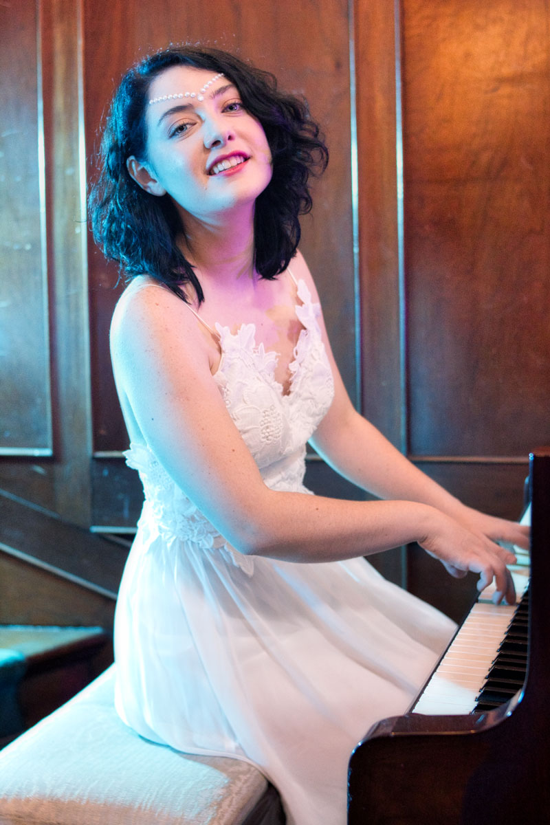 Retrato de Victoria Daninisa tocando piano con vestido blanco de encaje, sonriendo, sesión editorial en interior vintage CDMX, fotografía por Josué Palma